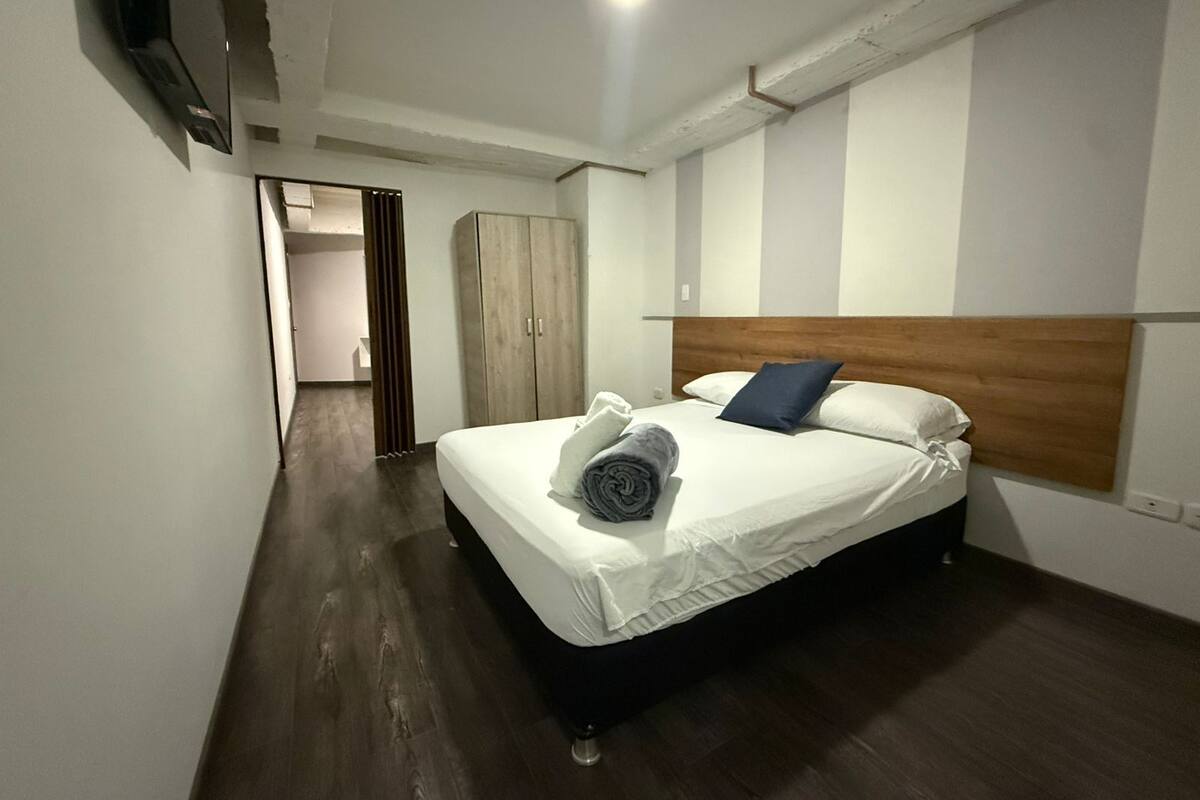 Habitación de hotel con una cama, cabecera de madera, armario y televisor montado en la pared.