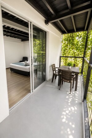 A balcony with a table and chairs, a sliding glass door leading to a bedroom with a bed and a chair, and a view of greenery. - Carriqui Underground  (Pereira)