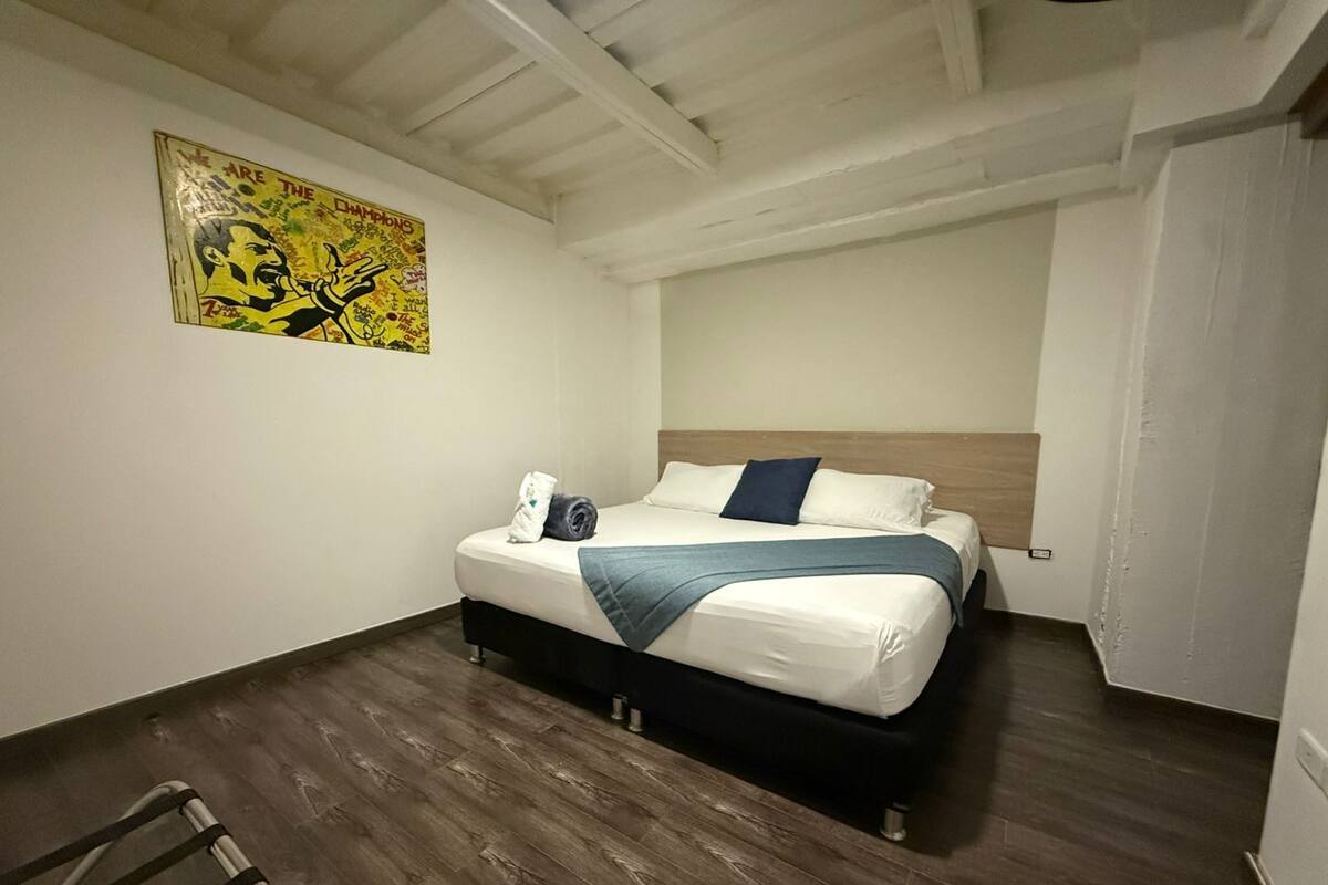 Una habitación de hotel con cama, suelo de madera, cabecero, toalla y un cuadro en la pared.