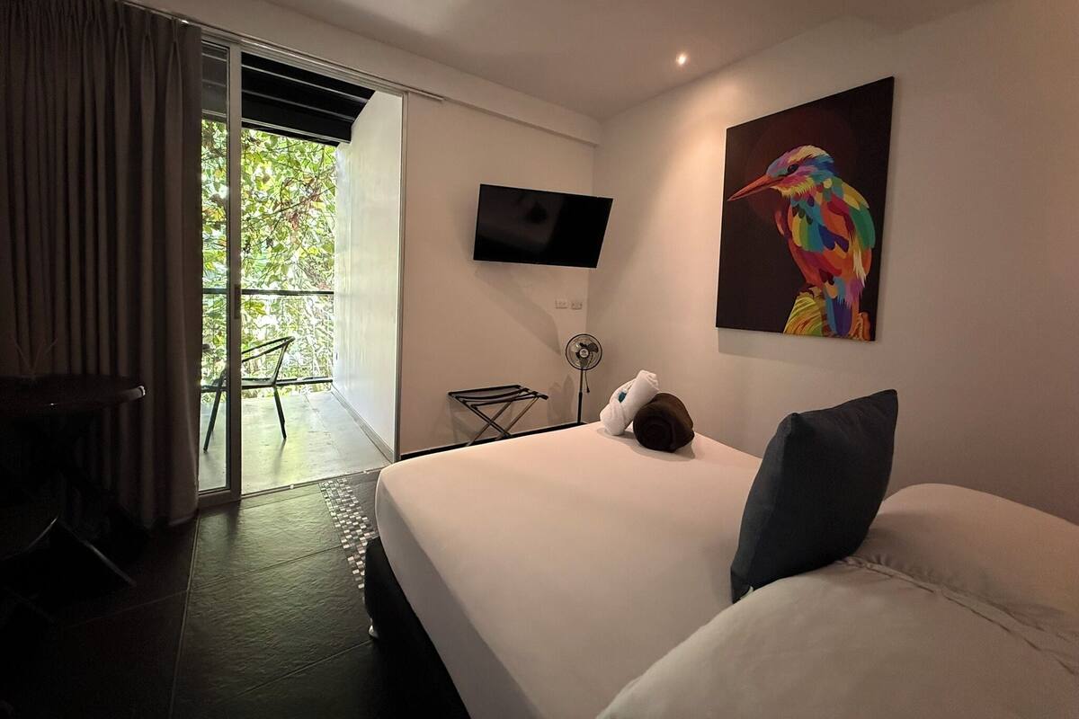 Habitación de hotel moderna con cama, televisor de pantalla plana, un cuadro, ventilador y vista a follaje por la ventana.