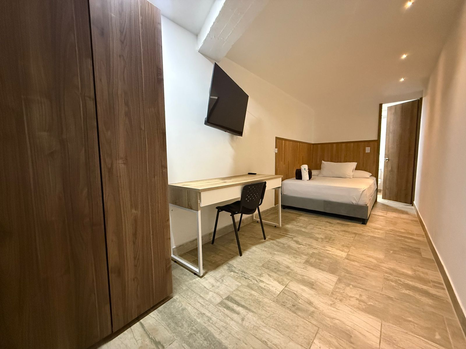 A modern hotel room with a wooden wardrobe, a desk with a chair, a bed, and a flat-screen TV mounted on the wall.