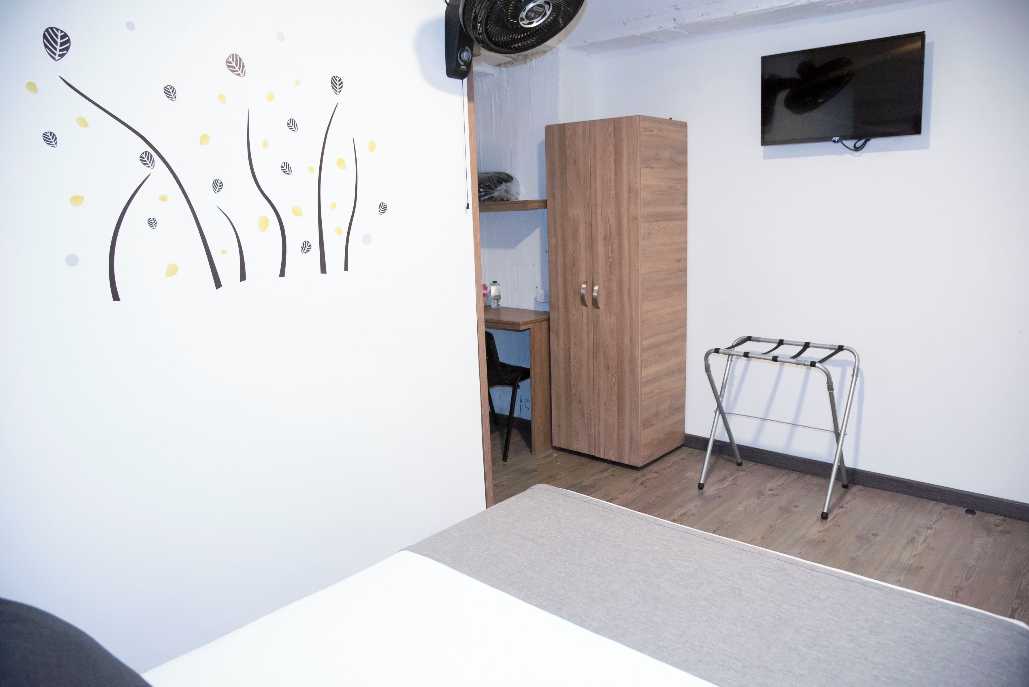 A room with a wooden wardrobe, a small desk, a TV mounted on the wall, and a drying rack.