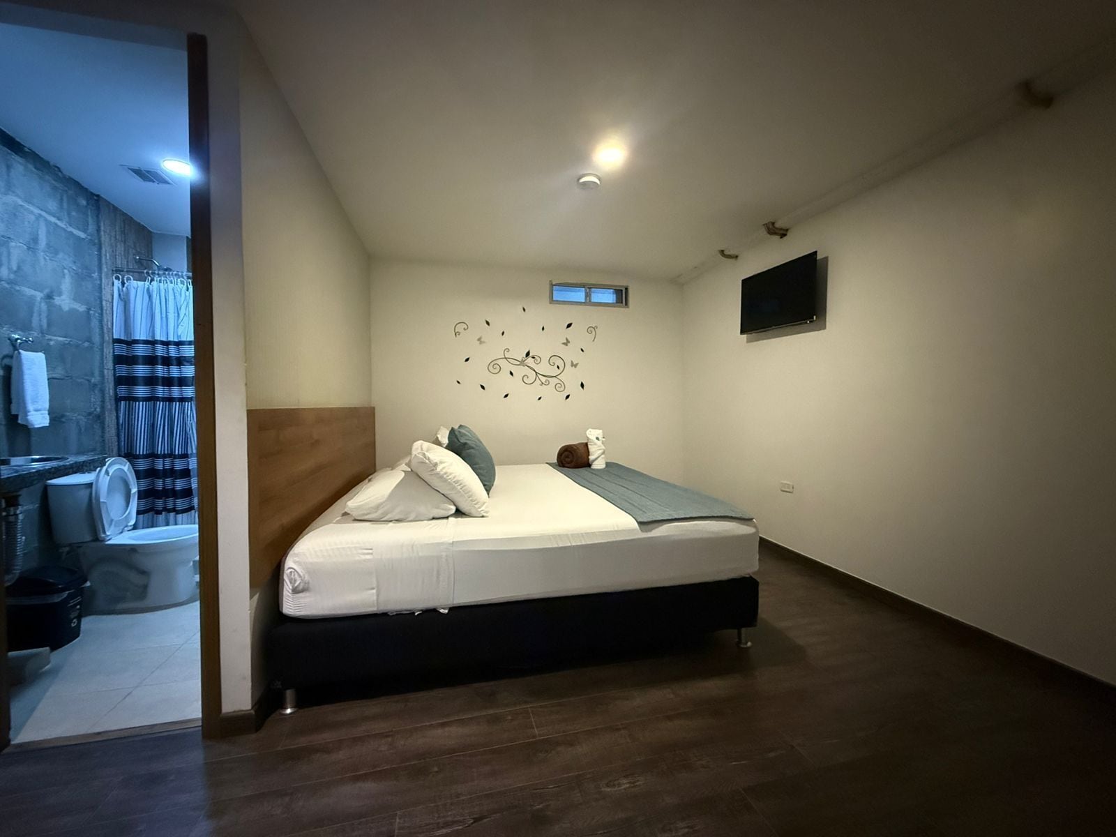 Une chambre moderne avec un lit, un téléviseur fixé au mur et une salle de bain visible à travers une porte ouverte.