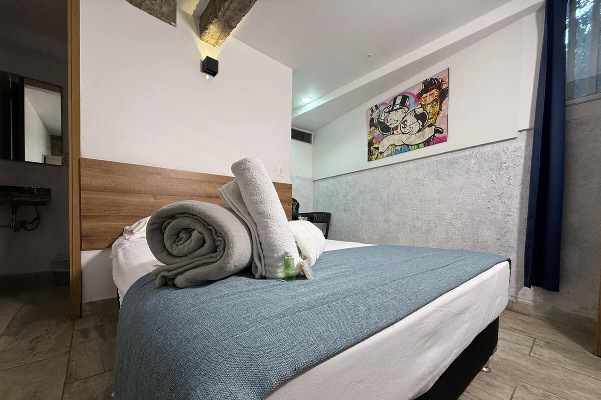 Una cama bien hecha con una manta azul y toallas enrolladas, un cabecero de madera, un cuadro en la pared y un baño con espejo y lavabo.