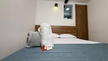 Una cama bien hecha con una manta gris y almohadas, una botella con líquido anaranjado y un paquete blanco etiquetado 'SENSY' sobre una colcha azul.