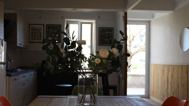A dining table with a vase of flowers, a kitchen in the background, and a window with natural light. - Provencal house with heated pool between Luberon & Alpilles - absolute calm (Cheval-Blanc)