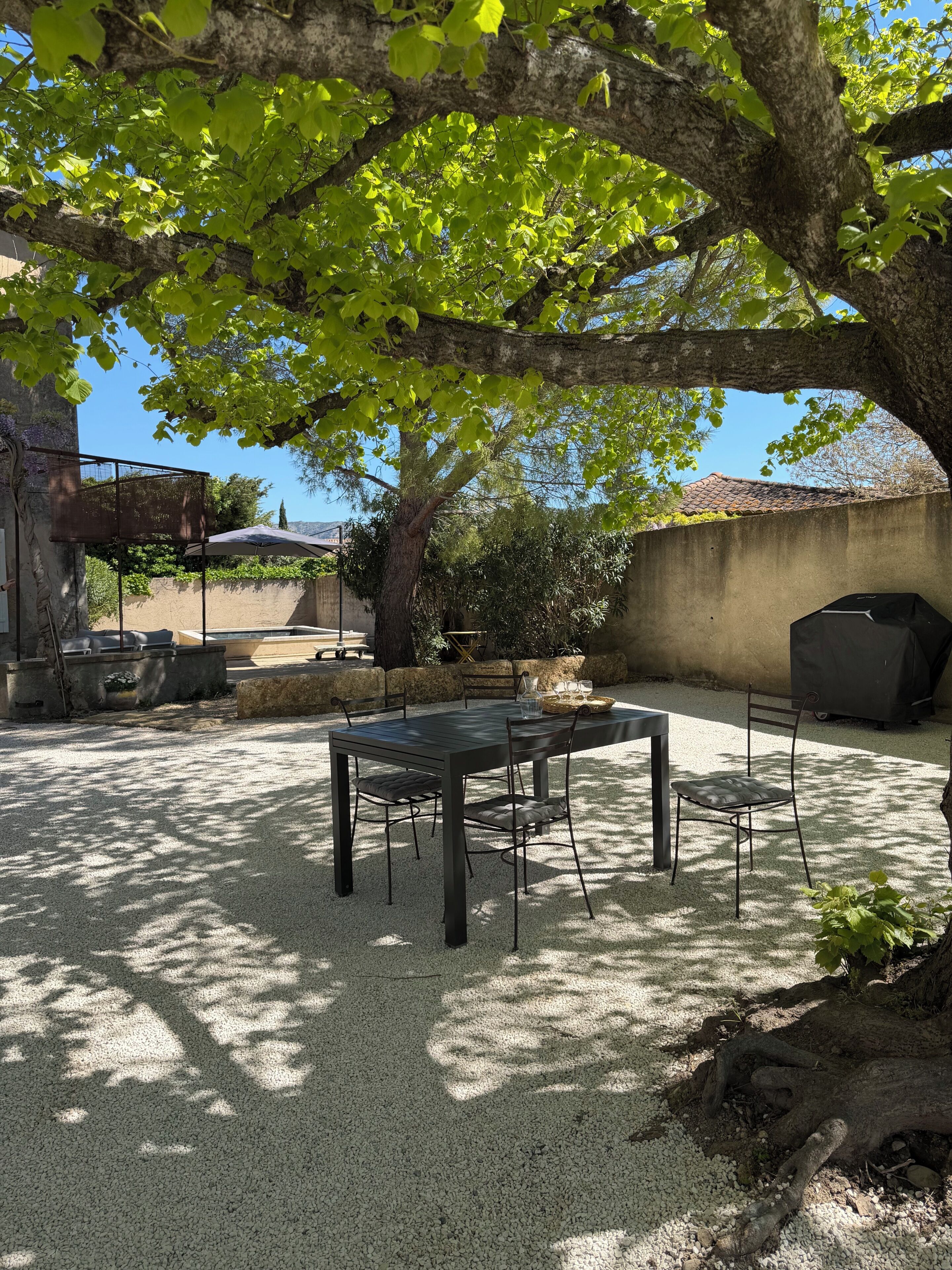 An outdoor patio with a table and chairs, surrounded by gravel and shaded by trees.
