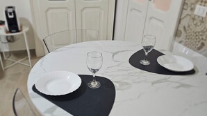 Une table à manger avec deux assiettes blanches et des verres à vin, des chaises transparentes et une machine à café en arrière-plan.