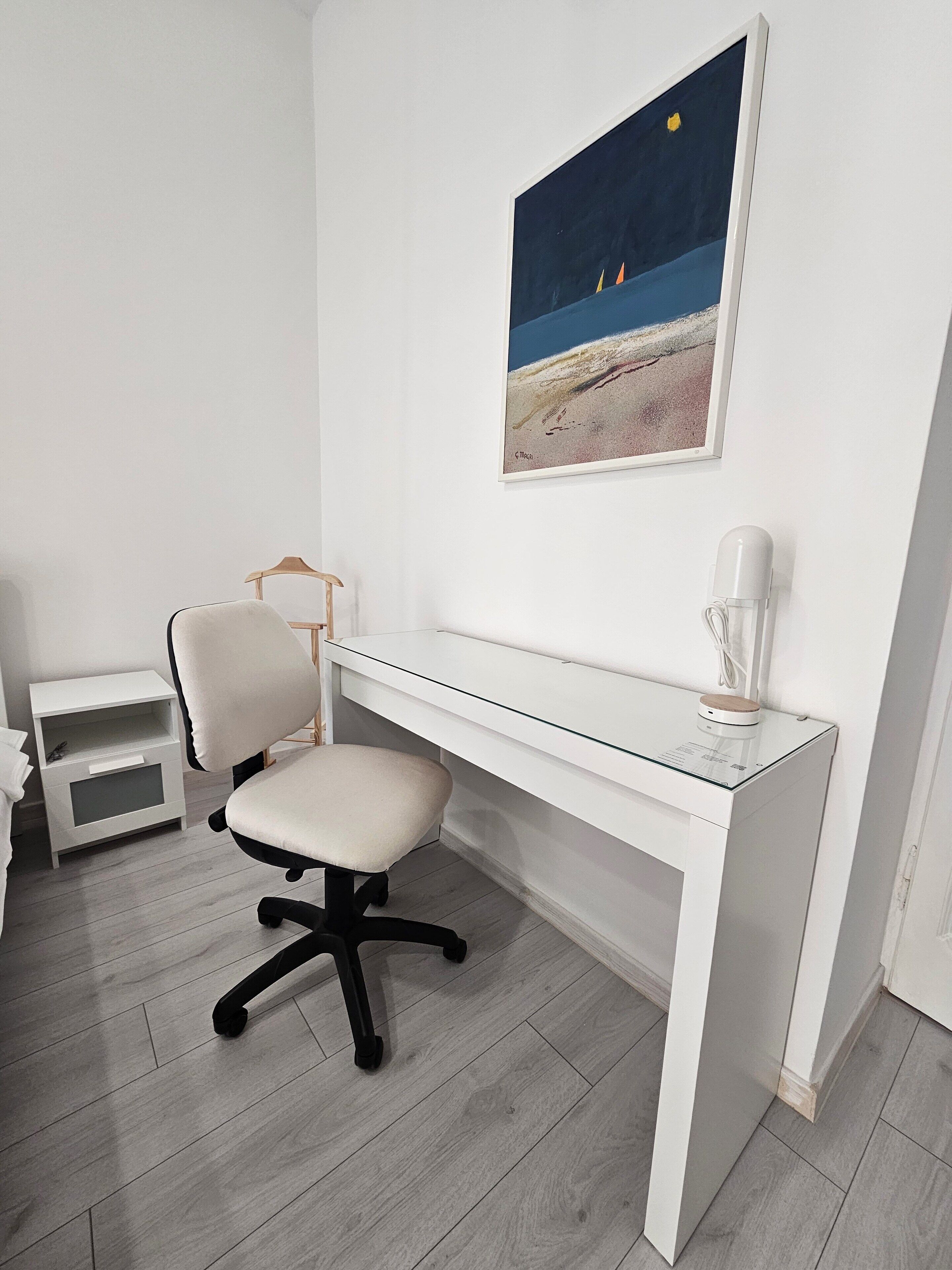 Un bureau blanc avec un plateau en verre, une chaise blanche, une petite table de chevet blanche et, au mur, un tableau représentant une scène de plage.