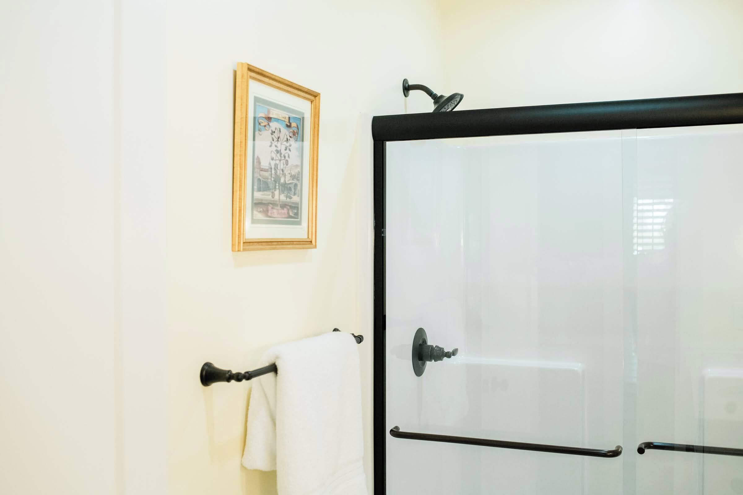 A bathroom with a glass shower door, a towel rack with a white towel, and a framed picture on the wall.