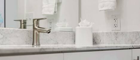 Une salle de bain moderne avec un comptoir en marbre, un lavabo avec un robinet chromé et un miroir au-dessus. On y trouve deux porte-serviettes avec des serviettes blanches et un rouleau de papier toilette sur le comptoir.