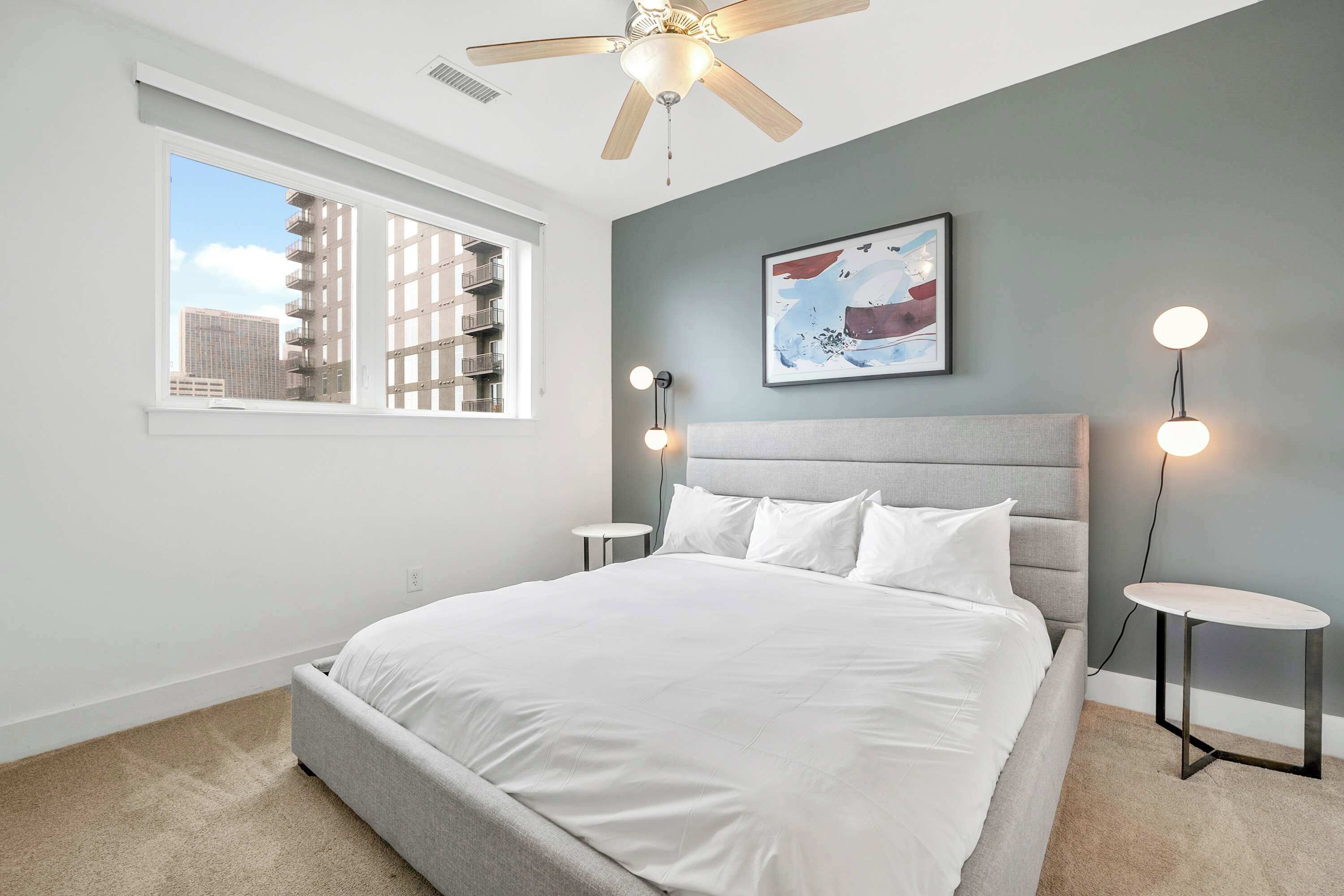 A bedroom with a bed, a ceiling fan, a window with a view of buildings, a wall-mounted light, and a small table.