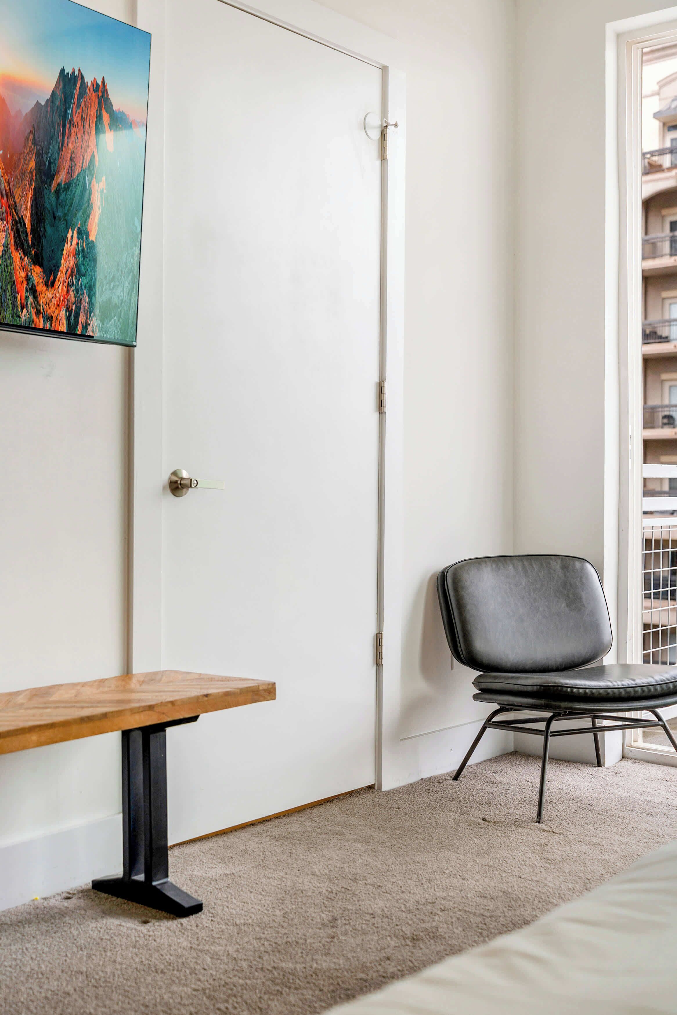 Una habitación con un banco de madera, una silla negra, una puerta blanca y un cuadro grande de una montaña.