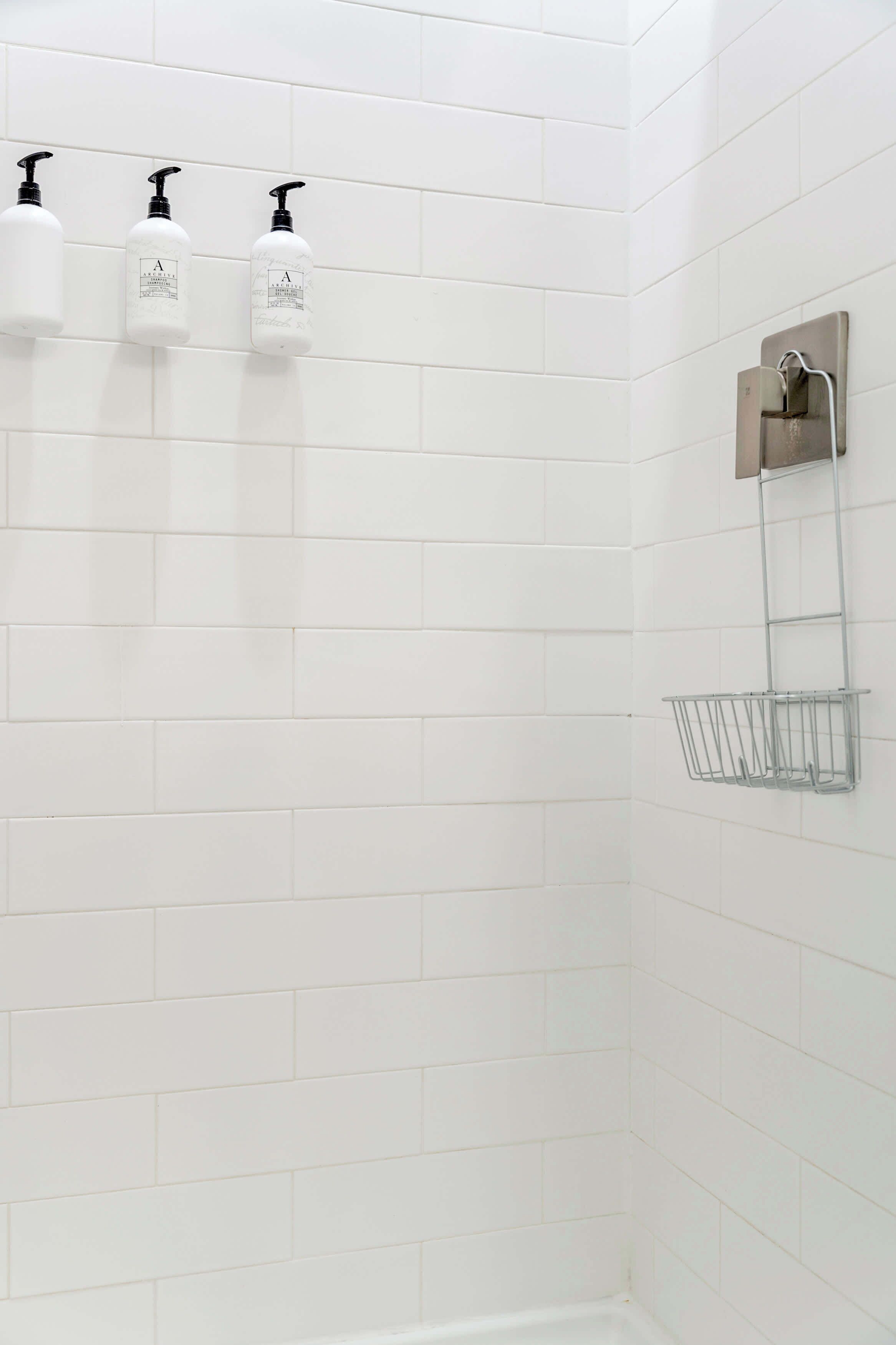 A bathroom with white tiled walls, three white bottles with black pumps, and a metal towel rack with a basket.