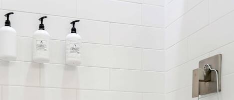 A bathroom with white tiled walls, three white bottles with black pumps, and a metal towel rack with a basket.
