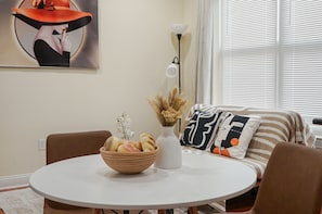 A dining area with a round table, a bowl of fruit, a vase with dried plants, and a modern painting on the wall.