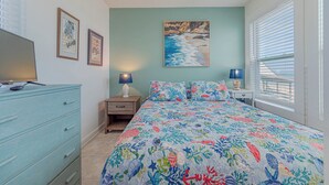 Een slaapkamer met een bed, een dressoir, nachtkastje, lamp, schilderij en jaloezieën.