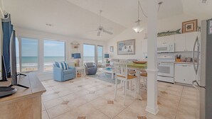 Een ruime woonkamer met uitzicht op het strand, een eethoek en een keuken met witte apparaten.