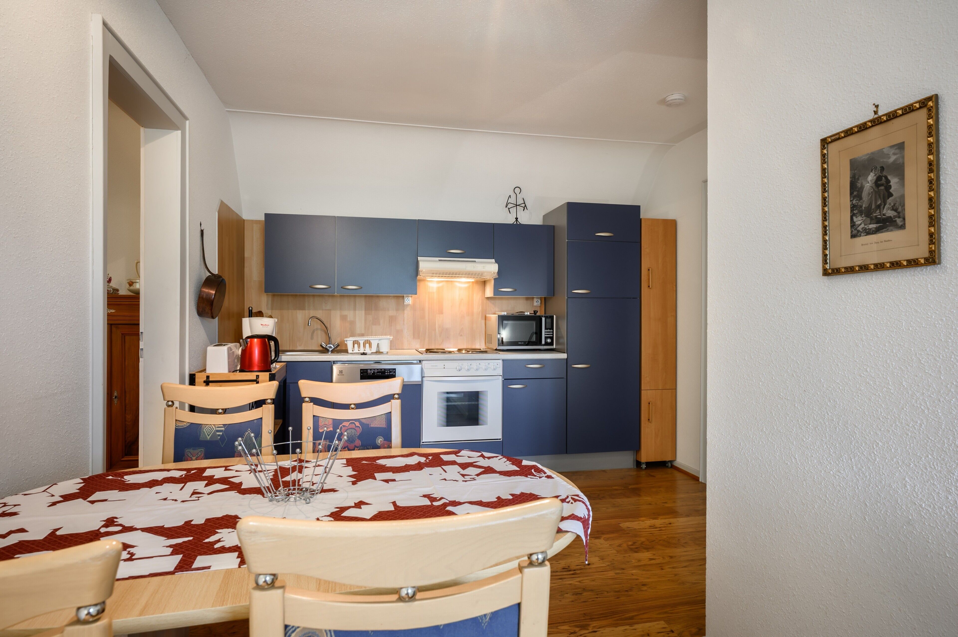 Een compacte keuken met blauwe kastjes, een witte kookplaat en een eethoek met tafel en stoelen.