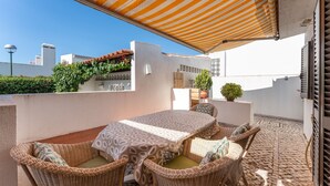 Un patio con mesa y sillas de mimbre, un toldo a rayas y plantas en macetas.