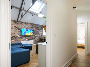 A modern living area with a brick wall, a flat-screen TV, blue sofas, and a wooden floor. - Serge | The Heim (Greater Manchester)