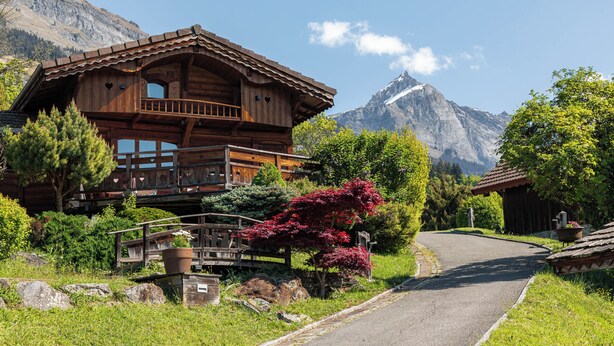 Eine Holzchalet mit einem Balkon, umgeben von üppigem Grün und Bergen im Hintergrund.