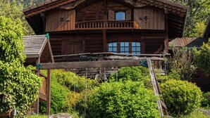 Eine Holzhütte mit Balkon, umgeben von üppiger Vegetation und Bergen im Hintergrund.
