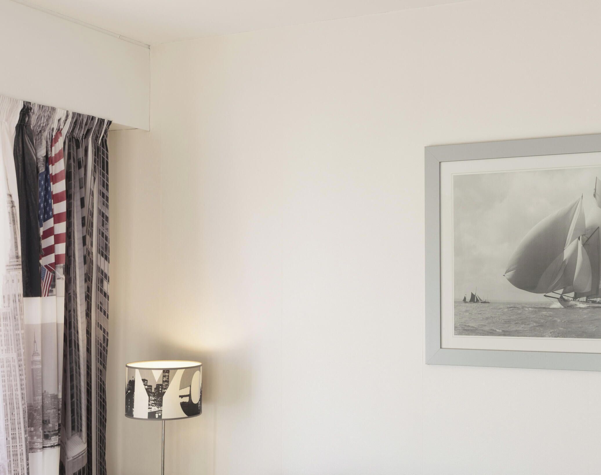 Una habitación con una fotografía en blanco y negro enmarcada de un velero en el agua, una lámpara con pantalla estampada y una cortina de rayas.