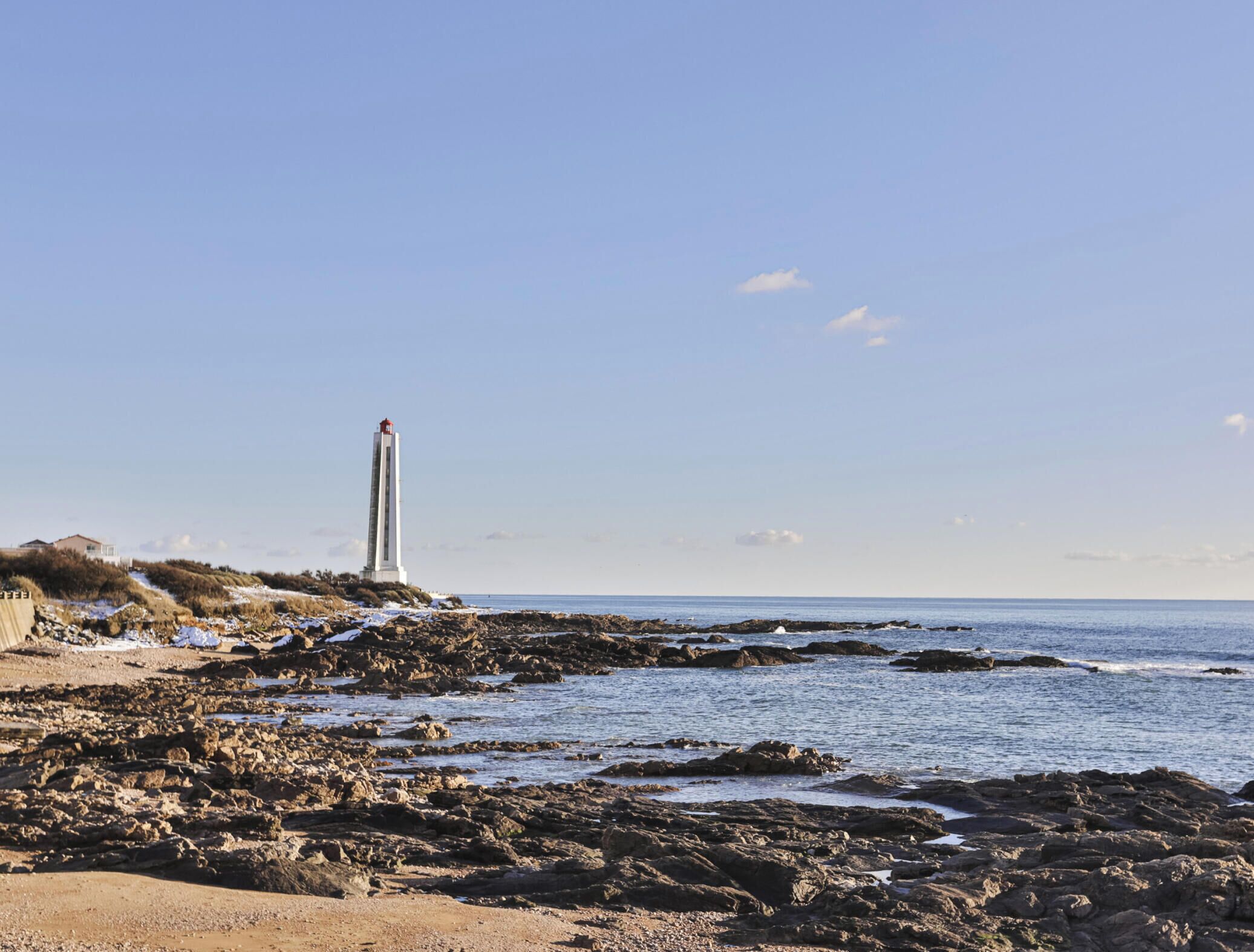Un paysage côtier avec un phare sur une côte rocheuse, une plage de sable et un ciel dégagé.
