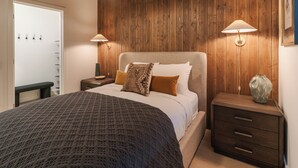 Quarto com cama grande, paredes de painel de madeira, duas luminárias de cabeceira, mesa de cabeceira e um banco.