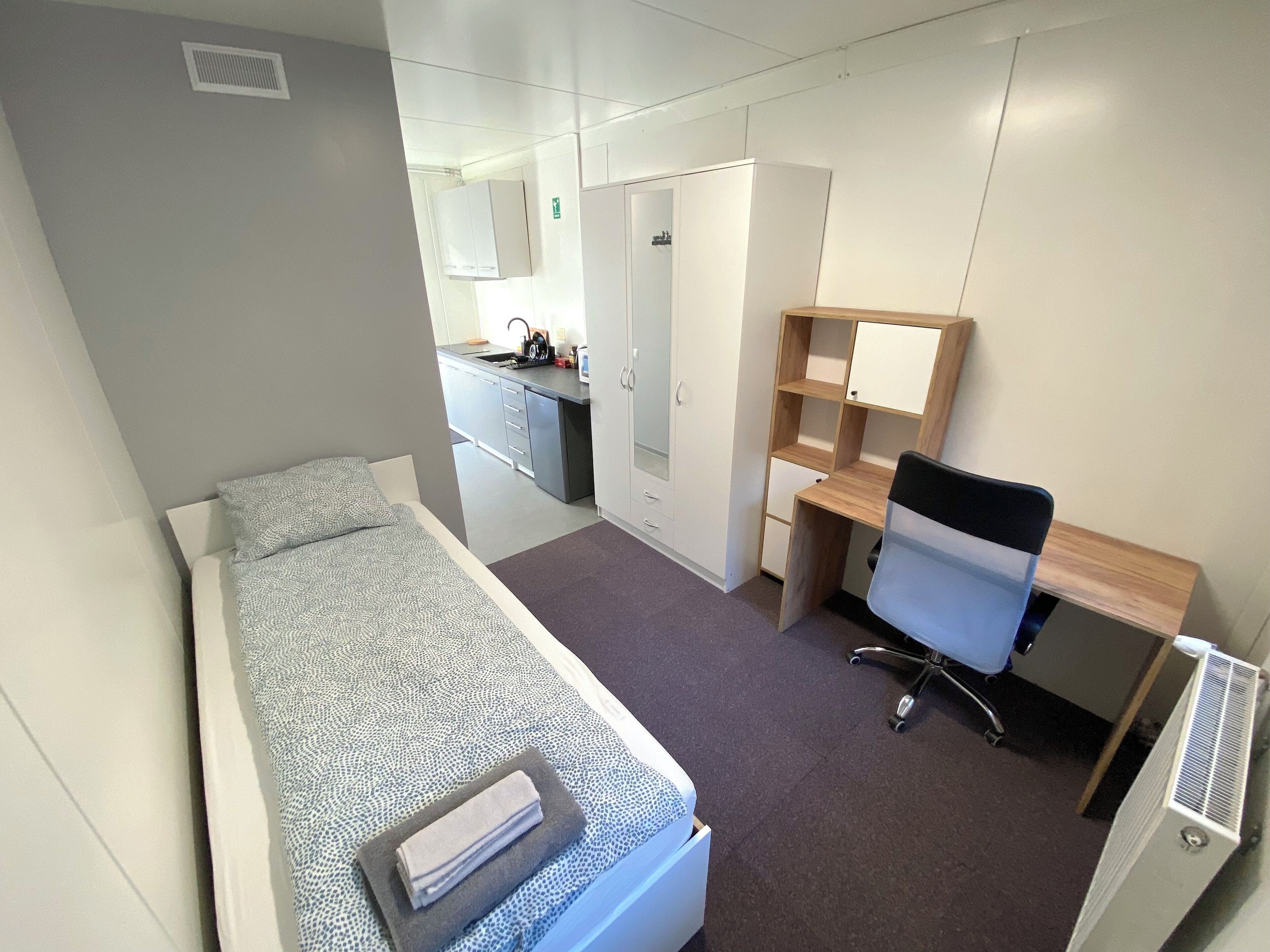 A compact room with a bed, a desk, a chair, and a kitchenette.