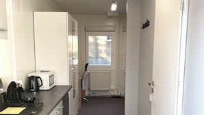 Kapea käytävä, jonka vasemmalla puolella on keittiöalue, jossa on mikroaaltouuni, vedenkeitin ja kahvinkeitin. Taustalla näkyy patteri, ja käytävä johtaa ovelle, jonka yläpuolella on kyltti.