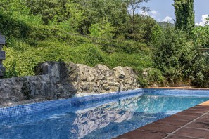 Piscina con baldosas azules, rodeada por un muro de piedra y abundante vegetación.