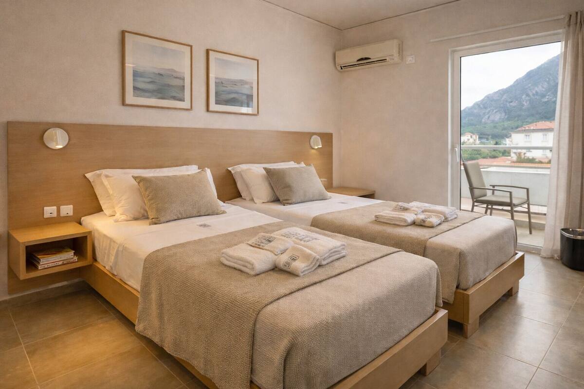 A hotel room with two beds, a small desk, a chair, and a balcony with a mountain view.