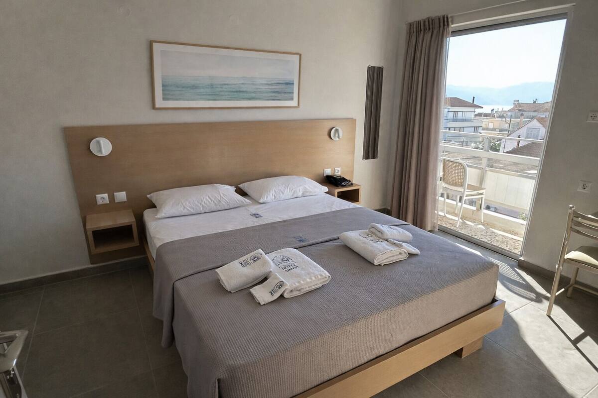 A hotel room with a bed, a bedside table, a nightstand, a window with a view, and a painting on the wall.