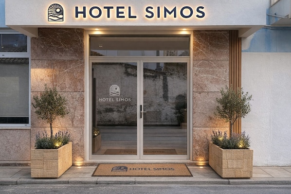 L’entrée de l’hôtel Simos présente un design moderne, avec une porte en verre, un paillasson et deux plantes en pot de chaque côté.