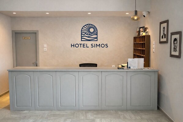 A hotel reception desk with a logo, a door labeled 'OFFICE', and a wall-mounted shelf with framed pictures. - Hotel Simos (Xiromero)