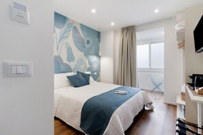 Ein Schlafzimmer mit Bett, fest installiertem Stromanschluss, Fenster mit Vorhängen, Schreibtisch mit Wasserkocher und Stuhl.