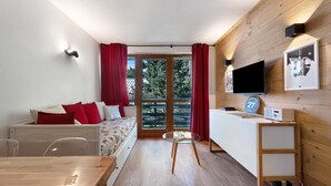A room with a wooden floor, a bed with red pillows, a glass table, a TV, and a window with curtains.