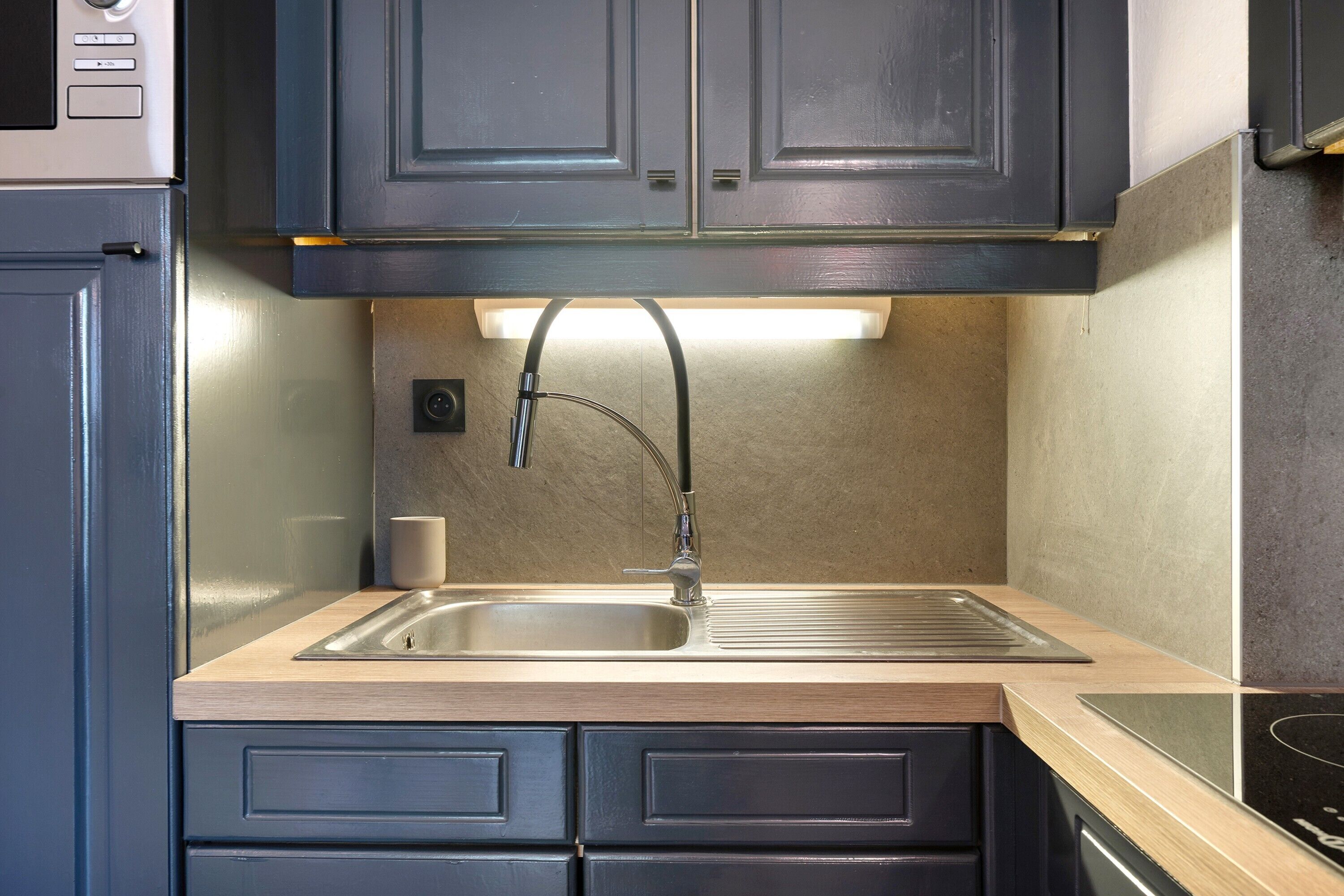 A kitchen with dark cabinets, a stainless steel sink, a built-in oven, and a light under the countertop.