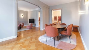 Ein moderner Essbereich mit einem runden Tisch und passenden Stühlen, ein Fernseher im Hintergrund und eine gemütliche Sitzecke mit Kissen.