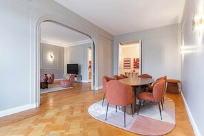 Ein moderner Essbereich mit einem runden Tisch und passenden Stühlen, ein Fernseher im Hintergrund und eine gemütliche Sitzecke mit Kissen.