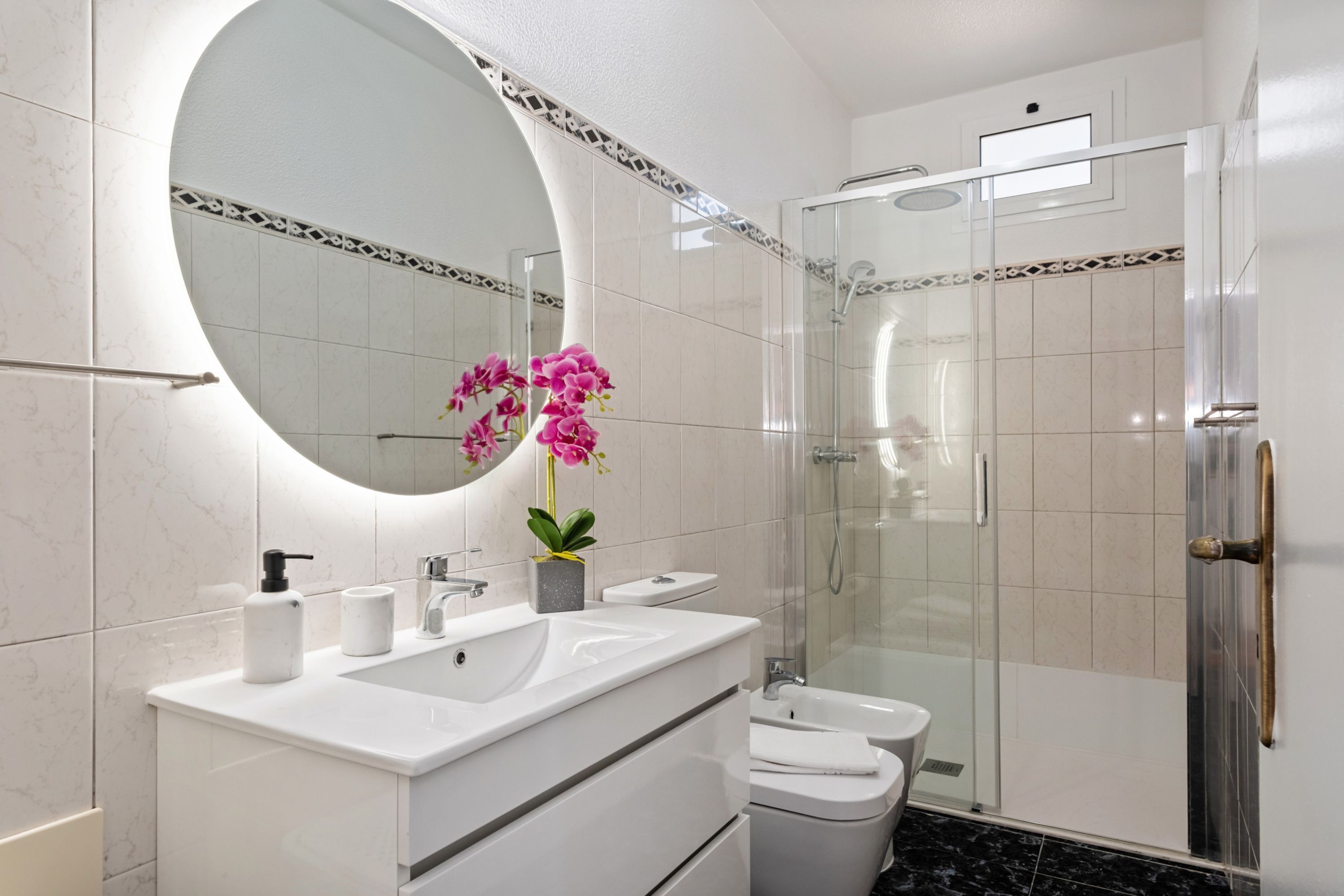 Ein modernes Badezimmer mit rundem Spiegel, weißem Waschtisch, Waschbecken und Toilette.