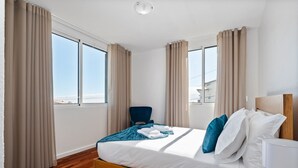 A hotel room with a wooden bed, beige curtains, a blue chair, and a view of the city. - John s Villa by Homie (Ponta do Sol)