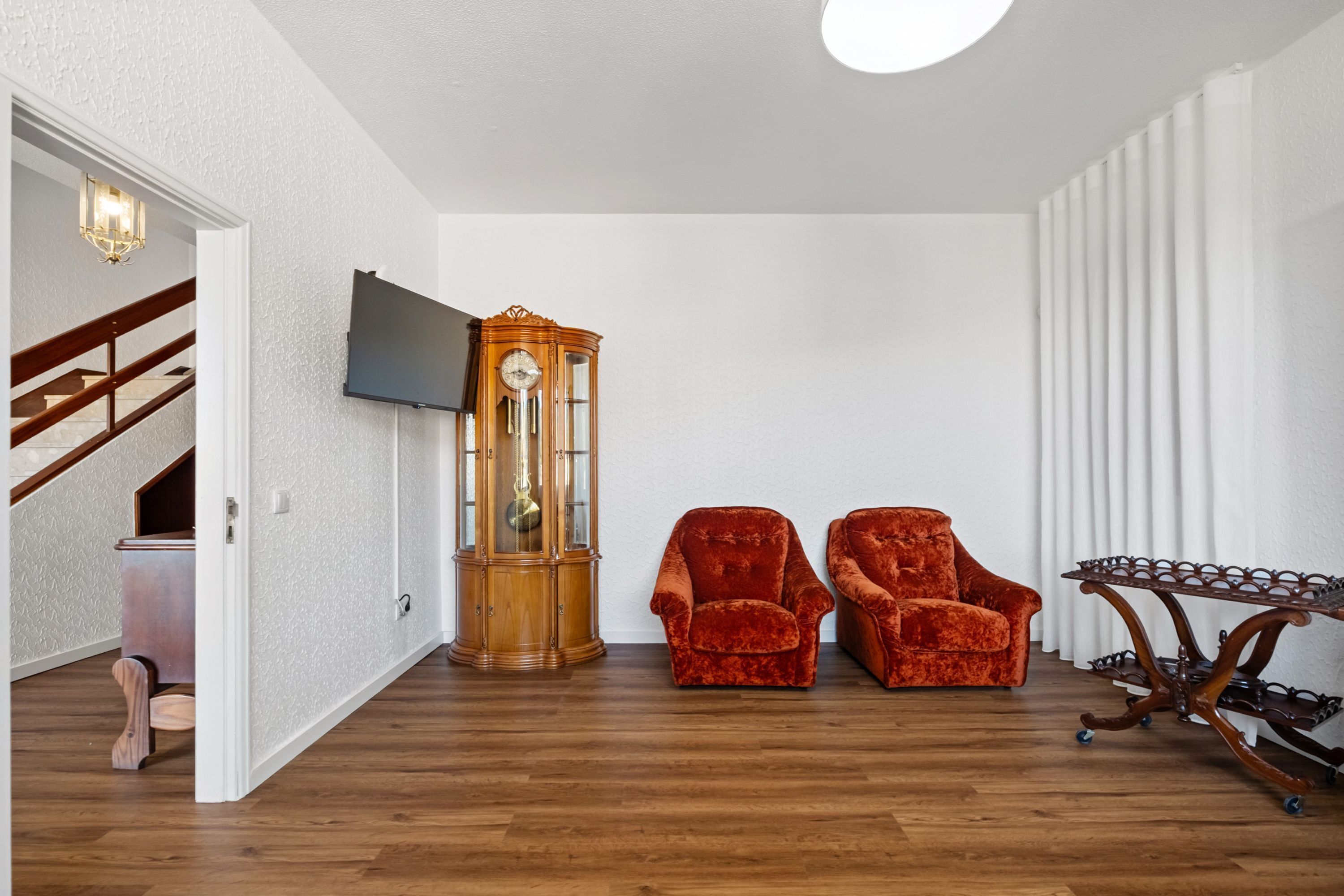 Una habitación con piso de madera, un reloj de madera grande, dos sillones de terciopelo rojo, una mesita de madera y un televisor montado en la pared.