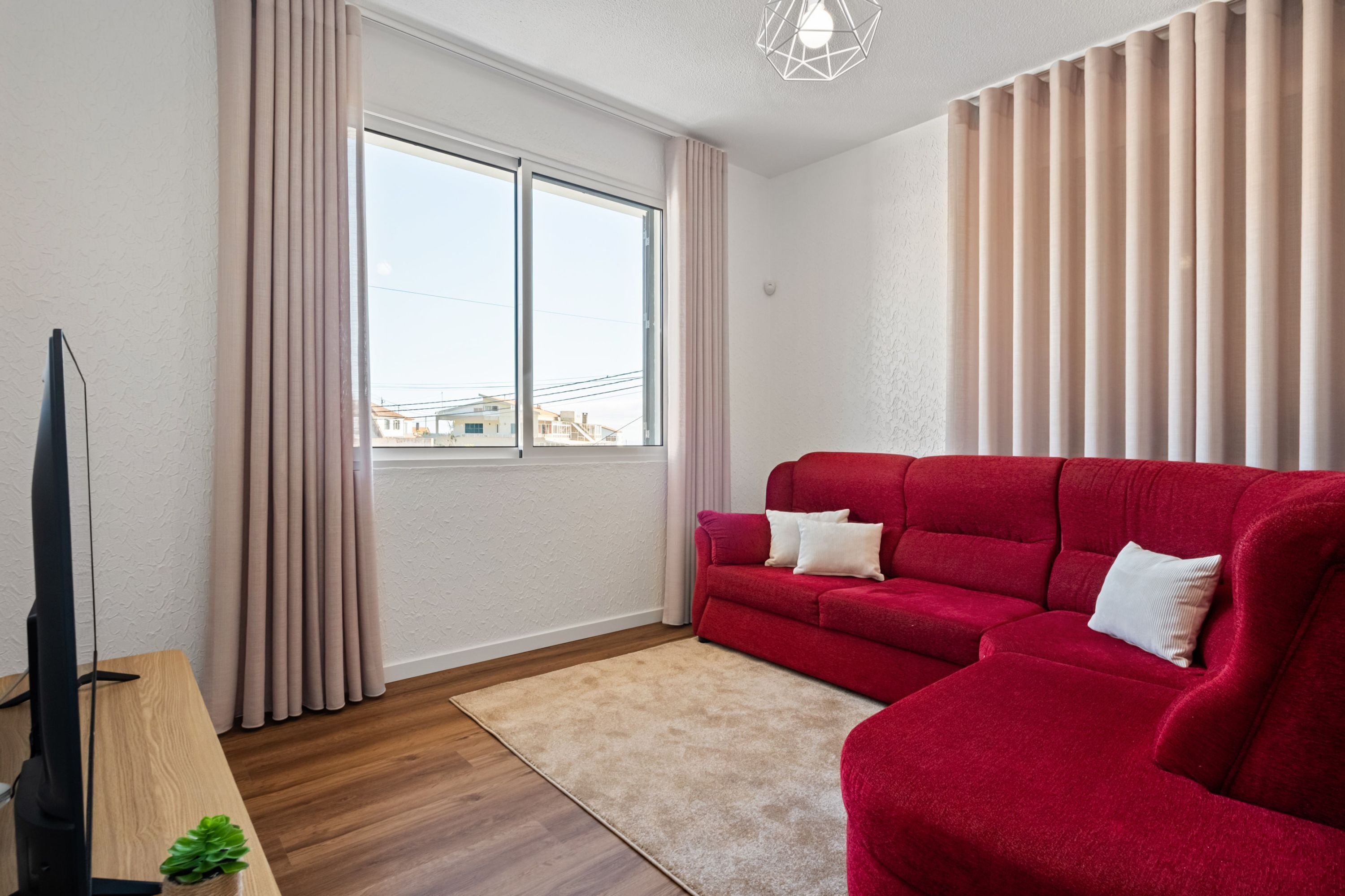 A modern living room with a red sofa, a flat-screen TV, and a large window with curtains.