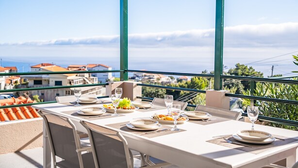 A balcony table set with plates, glasses, and a laptop, overlooking a coastal cityscape. - John s Villa by Homie (Ponta do Sol)