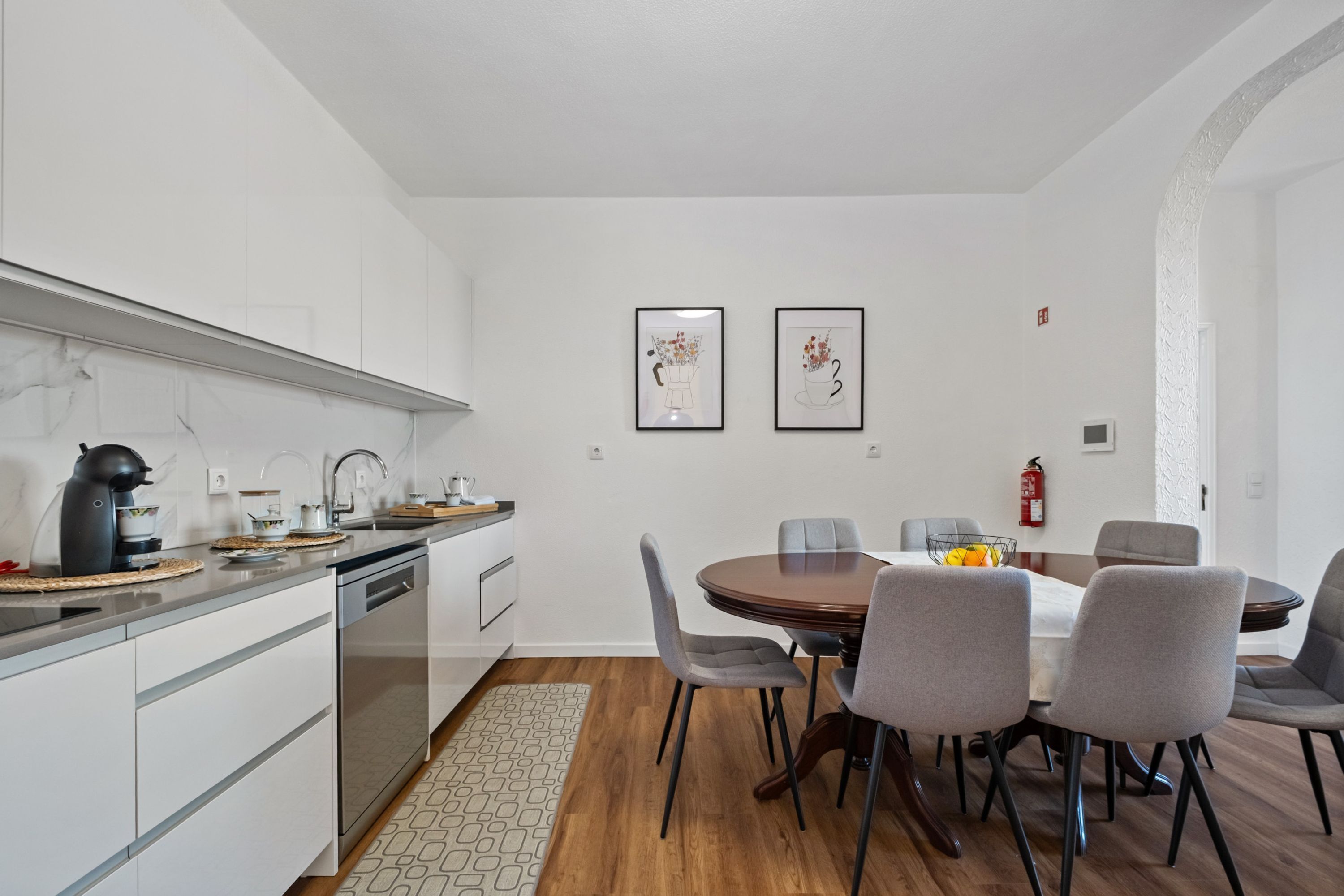 A modern kitchen with white cabinets, a built-in oven, and a coffee machine. There is a dining area with a round table and six chairs. Two framed pictures hang on the wall above the table.