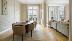 Ein Essbereich mit einem langen Tisch, Korbstühle, einer Couch mit Kissen und großen Fenstern mit Blick auf Gebäude.