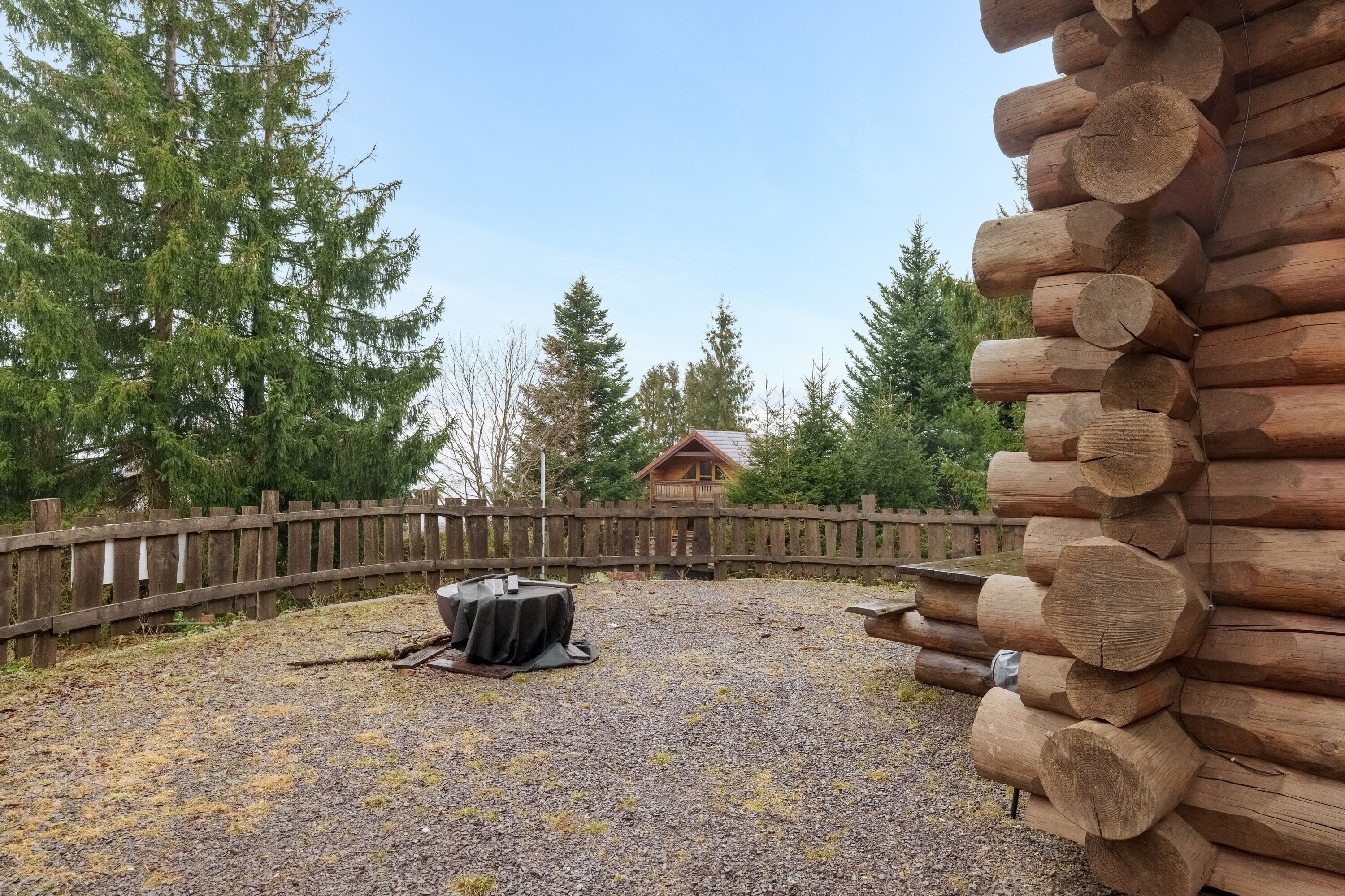 A log cabin with a gravel patio, a wooden fence, and a view of a cabin in the background.